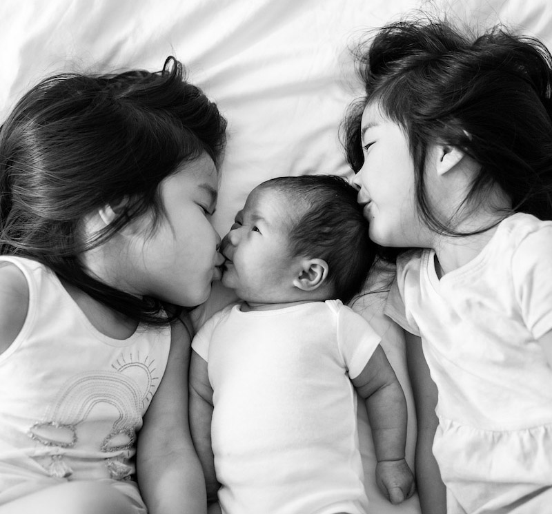 BW Sisters laying on a bed kissing their new baby sibling