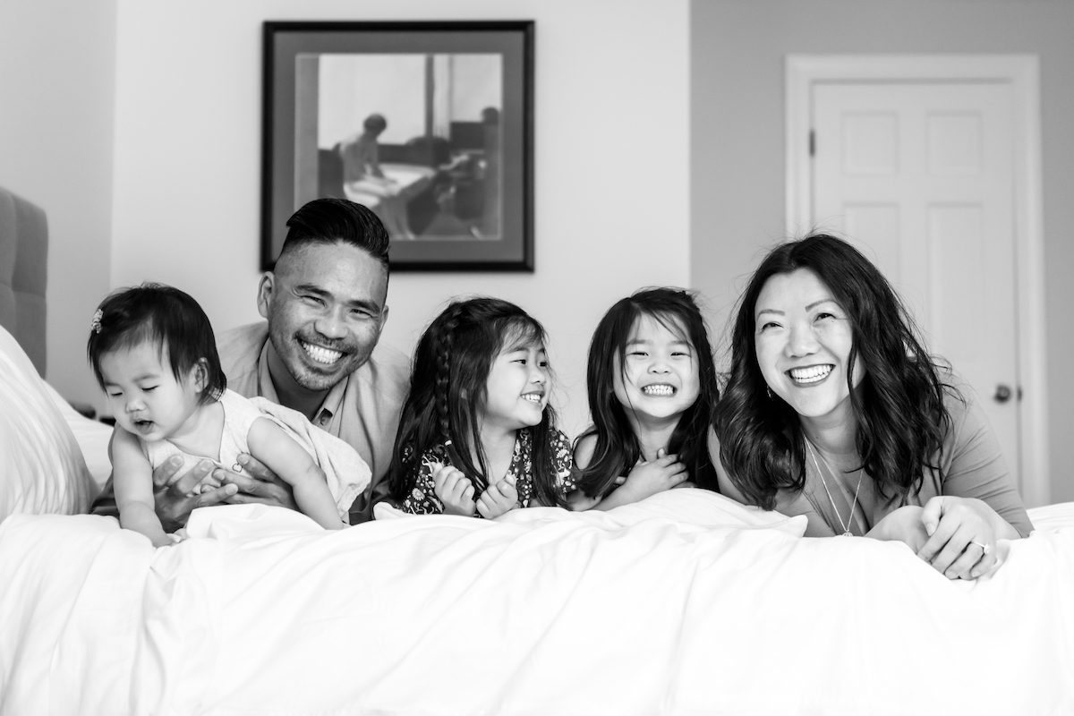 Parents laying on a bed with their three daughters lauging at the camera