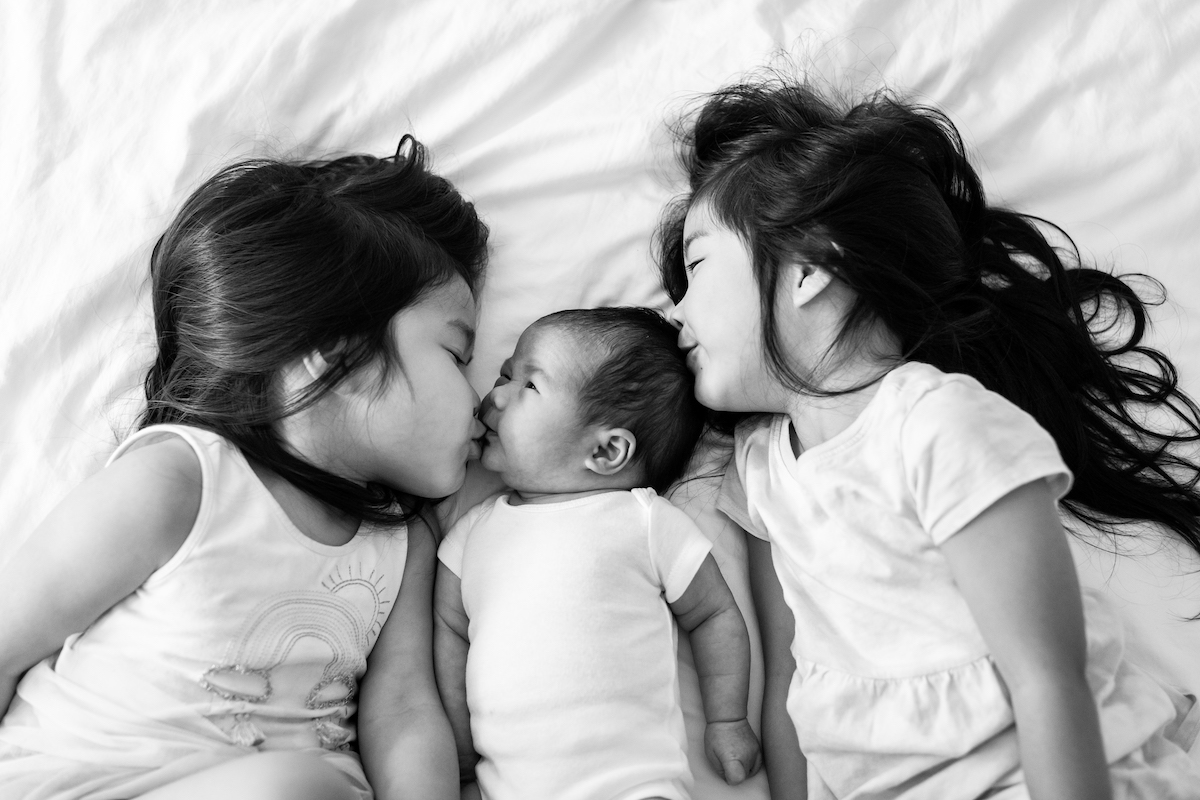 Sisters laying on a bed kissing their new baby sibling