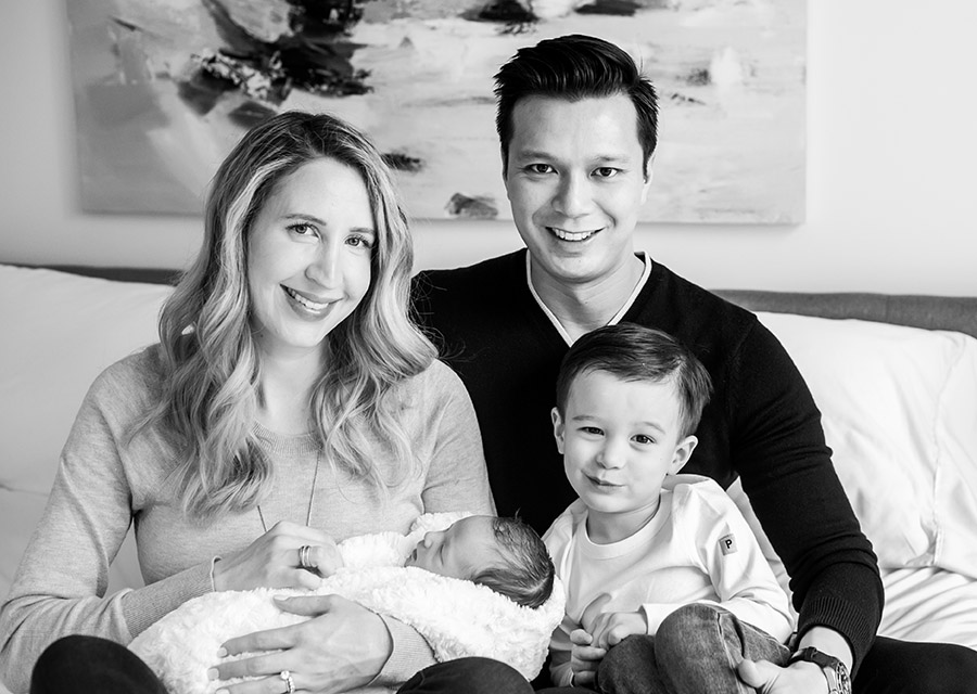 BW Parents with their toddler and their newborn baby on a bed smiling at the camera