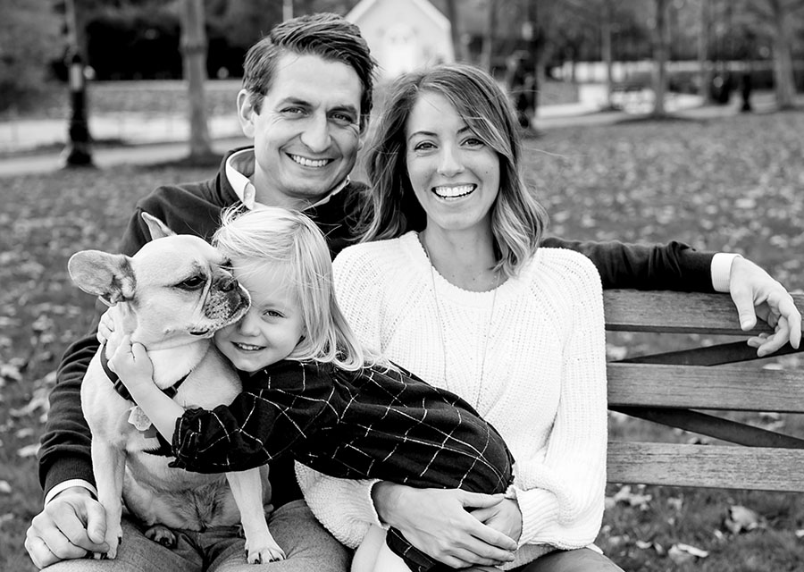 BW Family sitting on a bench in a park with their daughter and their dog smiling at the camera