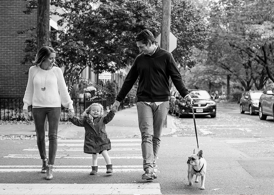 Parents holding their daughters hands while chossing a street with their dog