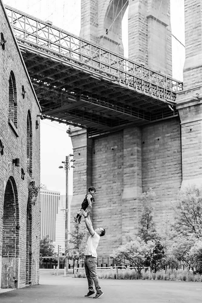BW Daddy throwing his toddler son into the air below brooklyn bridge in dumbo