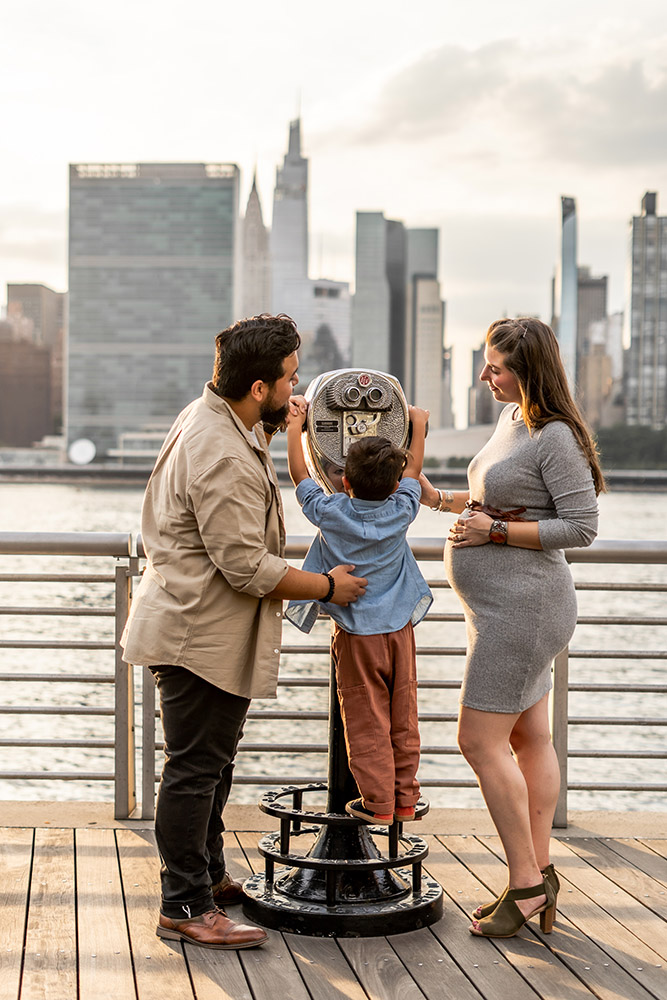 Beautiful woman showing off her belly with Manhattan skyline in the background, son trying to reach the binocular machine, dad helps him