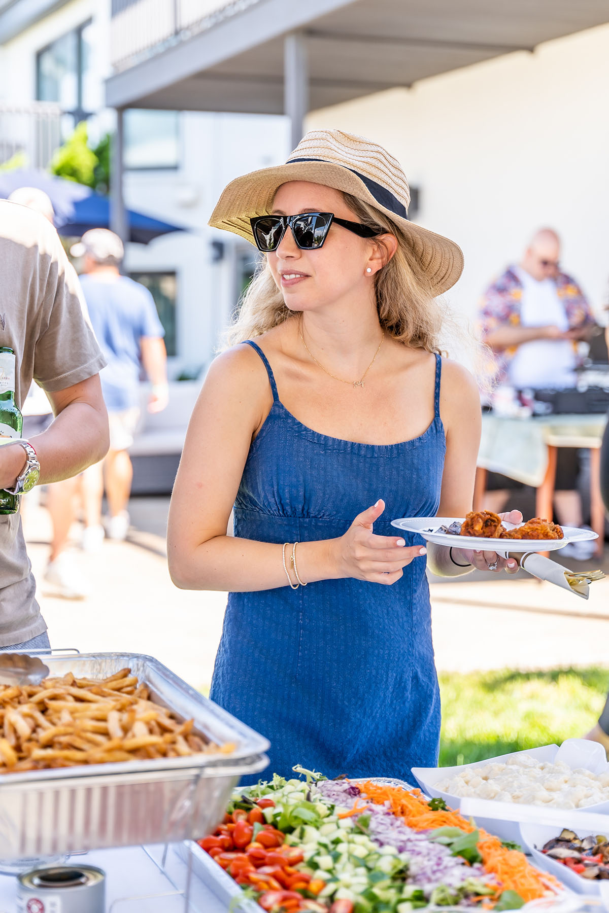 woman in blue dress with sunglasses and hat is holding food on a plate