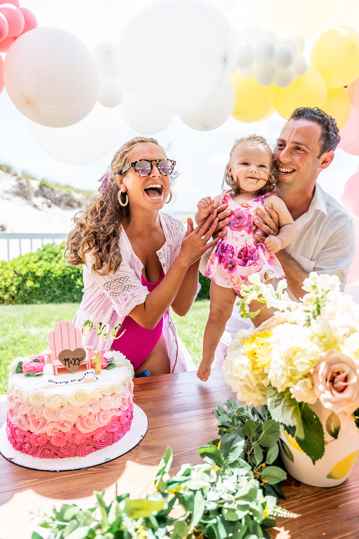 family of three laughing and sitting at a table with birthday cake and flowers balloons in background