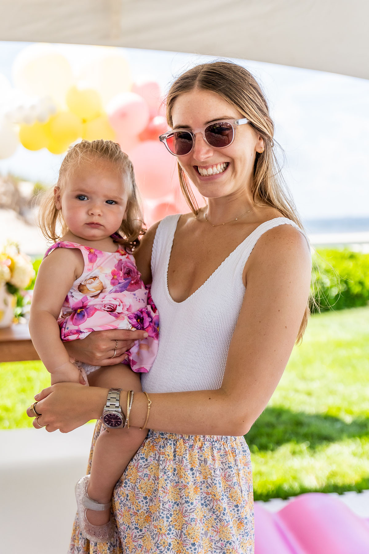 mom with sunglasses is holding a toddler in pink flower dress