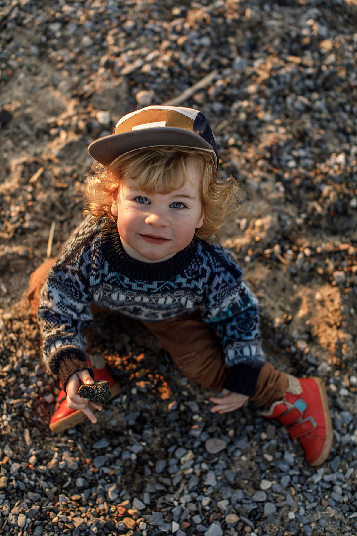 blond toddler with cap sitting on the ground looking up in the camera