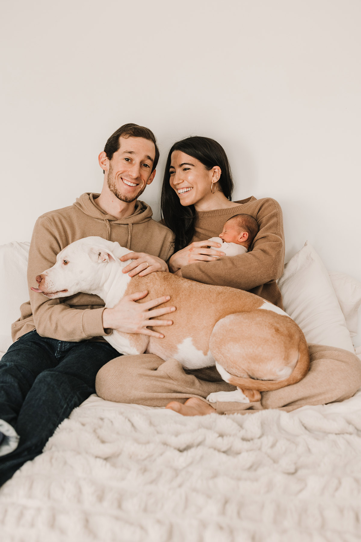 happy family of three with dog sitting on bed mom is holding their newborn