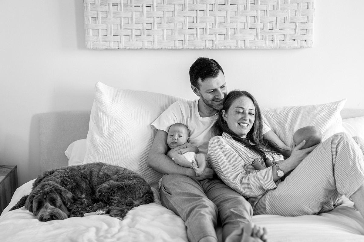 BW family with dog laying in bed mom and dad holding their newborn babies