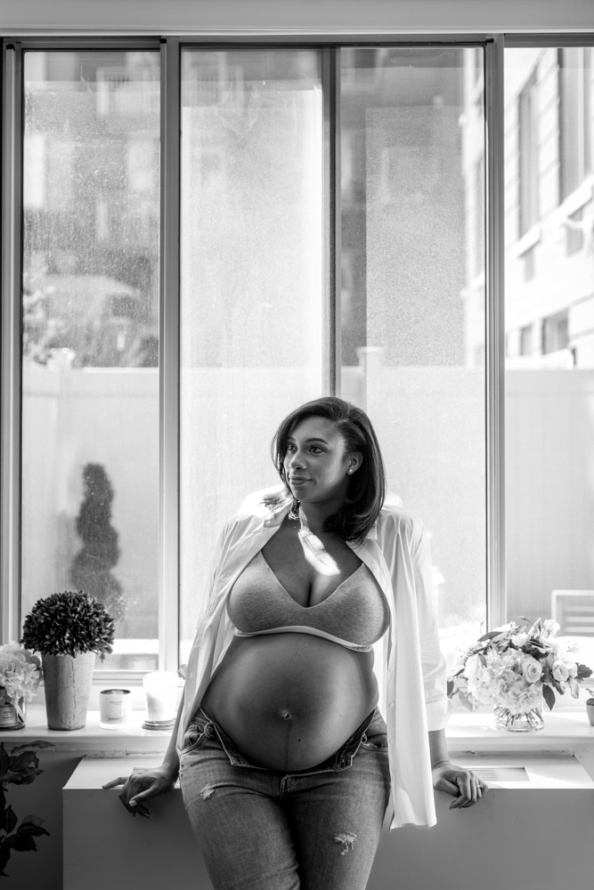 black and white photo of a pregnant woman leaning against a table showing her pregnant belly in opend jeans a bra and an open shirt