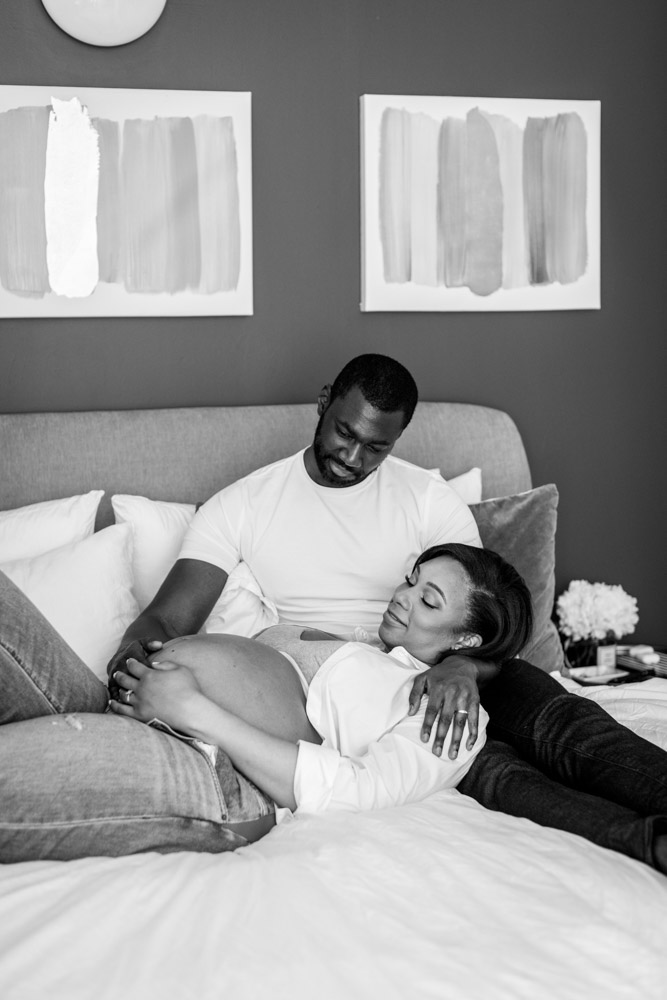 black and white photo of a couple lying on a bed both happily looking at the wife's pregnant belly