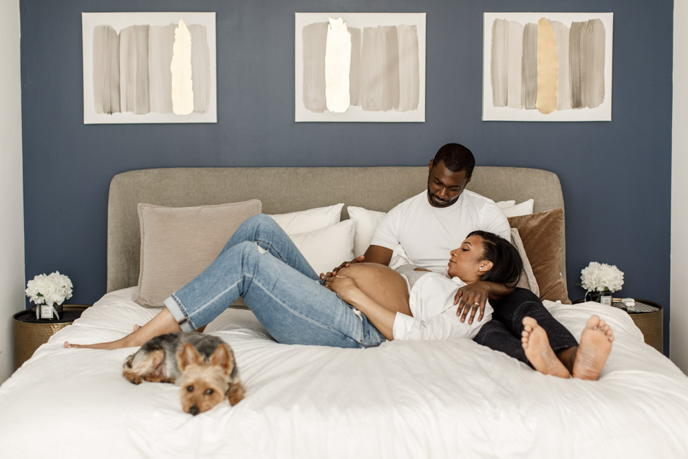 couple lying on a bed with their dog both have their hands on the wife's pregnant belly