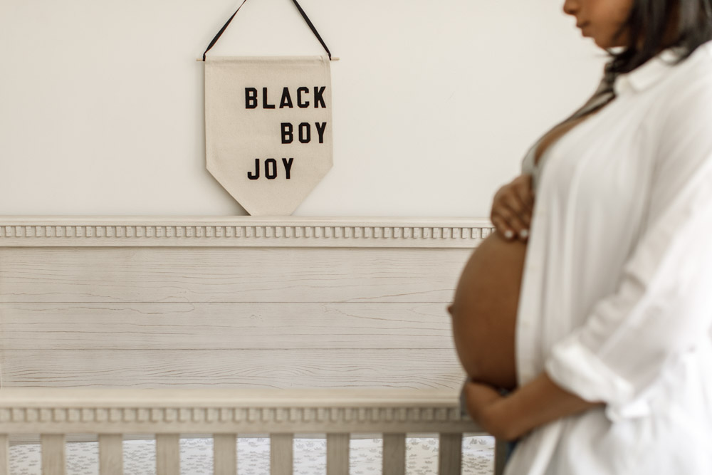 pregnant woman standing in front of a crib embracing her pregnant belly, focus is on the sign hanging above the crib that says "black boy joy" in capital letters