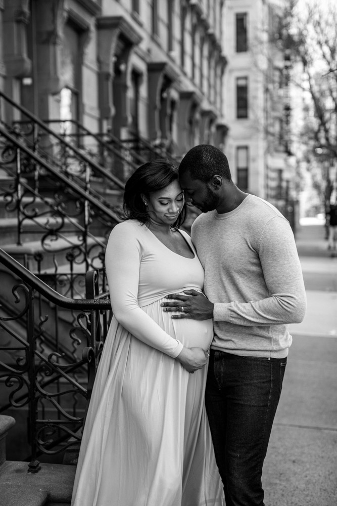 black and white photo of a couple standing on a sidewalk, heads close together, the man has his hand on his wife's pregnant belly