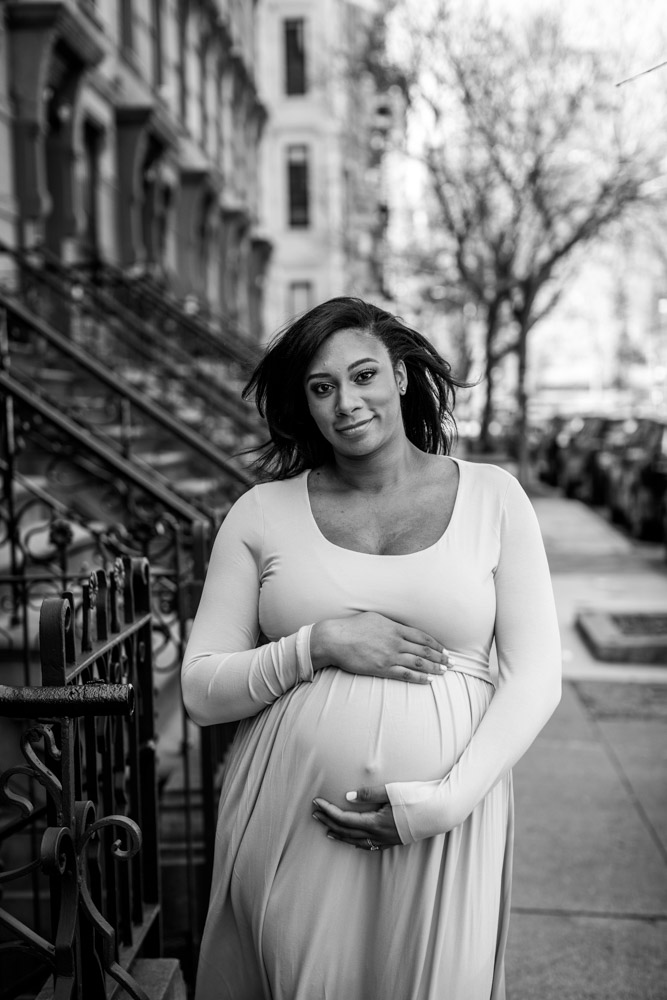 black and white photo of a woman standing on a sidewalk smiling at the camera as she is embracing her pregnant belly