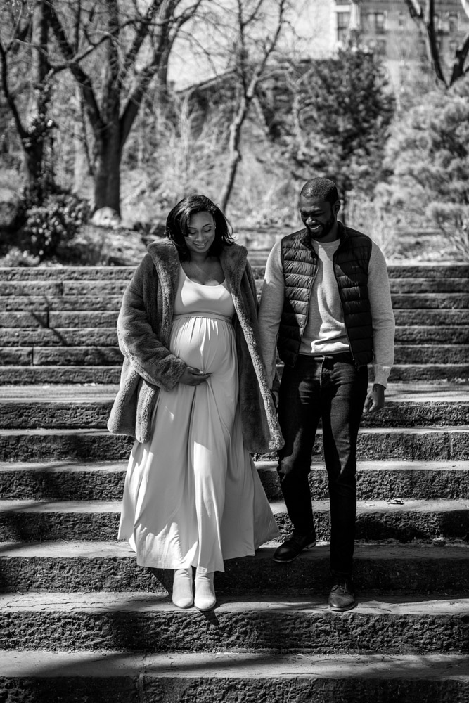 black and white photo of a couple laughing as they walk down some steps in a park holding hands the woman is holding her pregnant belly