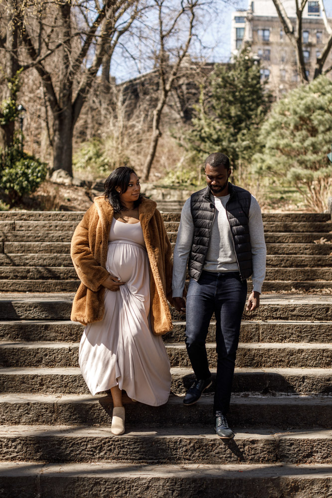 couple walking down some steps in a park holding hands the woman is holding her pregnant belly