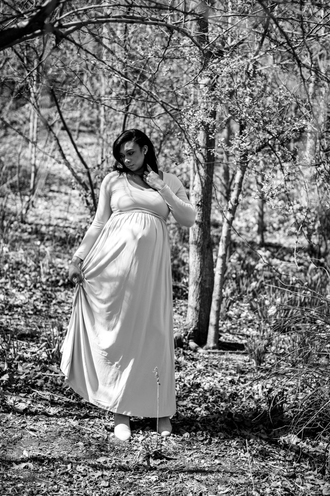 black and white photo of a pregnant woman standing in the woods in a long dress facing the camera holding on to her dress with one hand and brushing trough her hair with the other