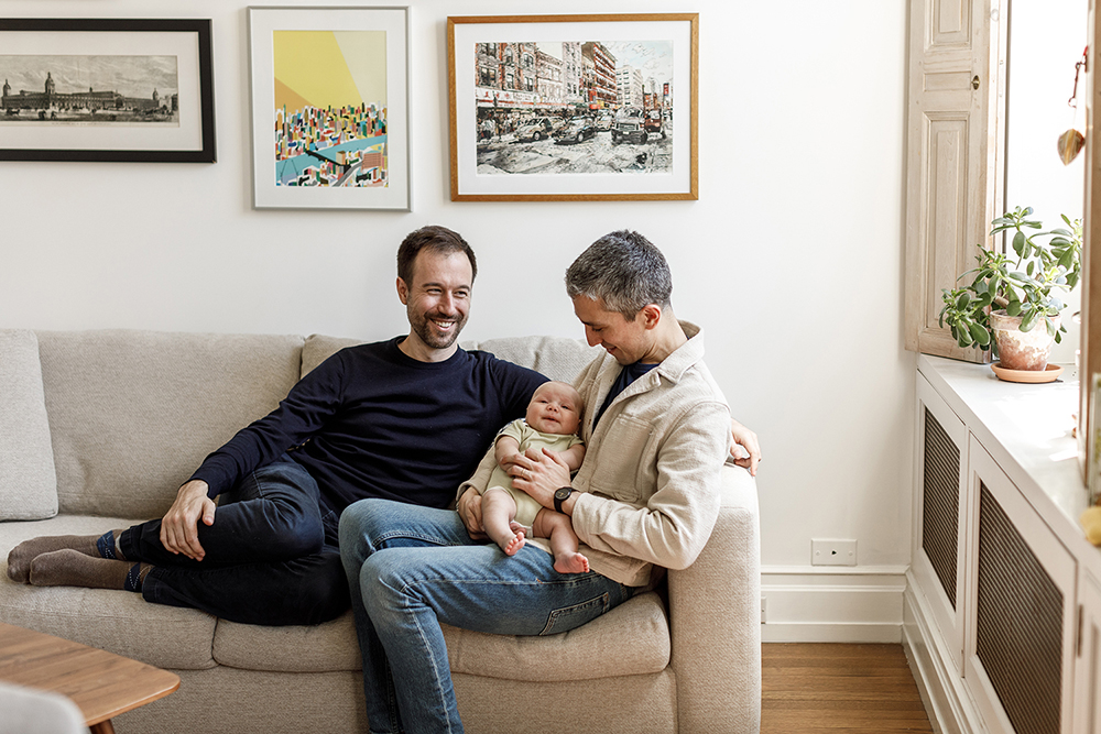 couple sitting on a couch one dad is holding their baby smiling while the other dad is smiling at his partner