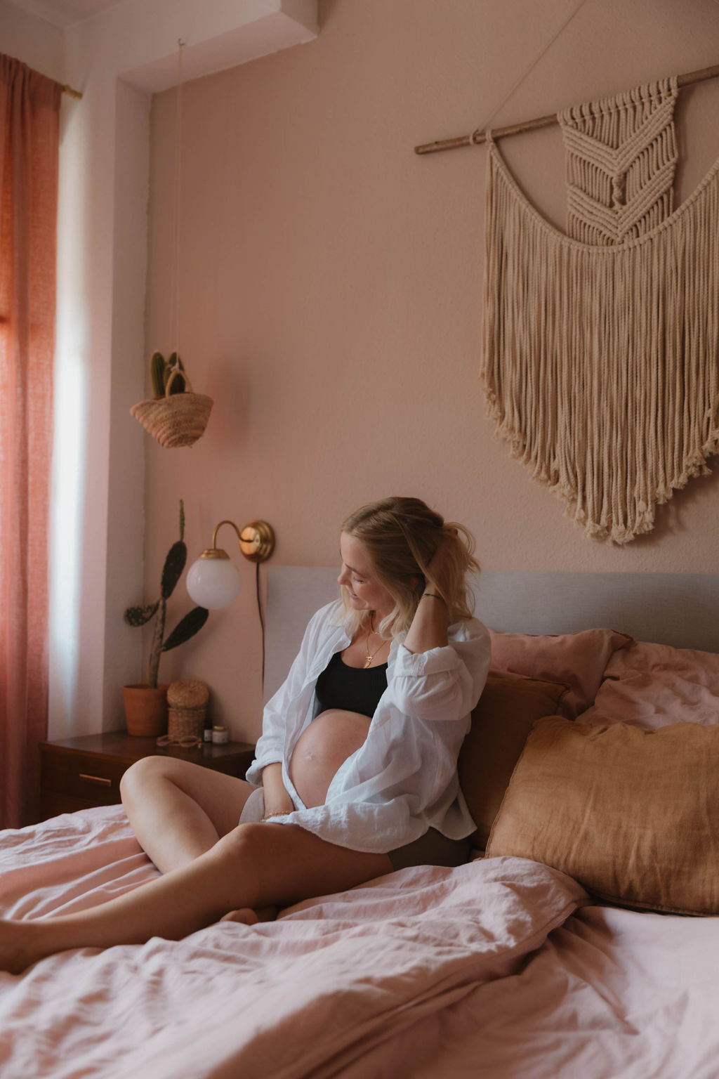 pregnant woman sitting on a bed wearing shorts a bra and an open shirt showing her pregnant belly
