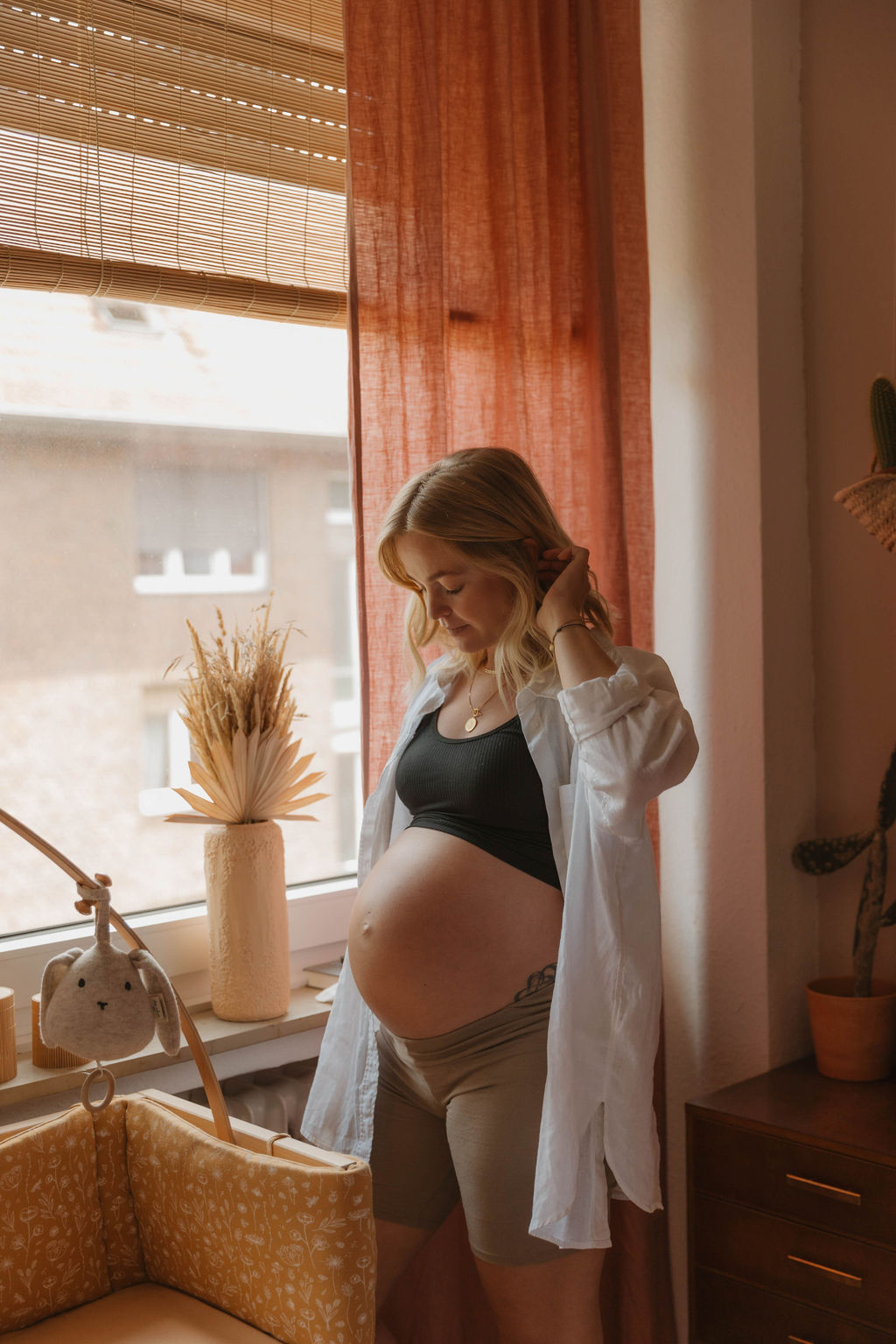 pregnant woman standing in front of a big window wearing shorts a bra and an open shirt showing her pregnant belly