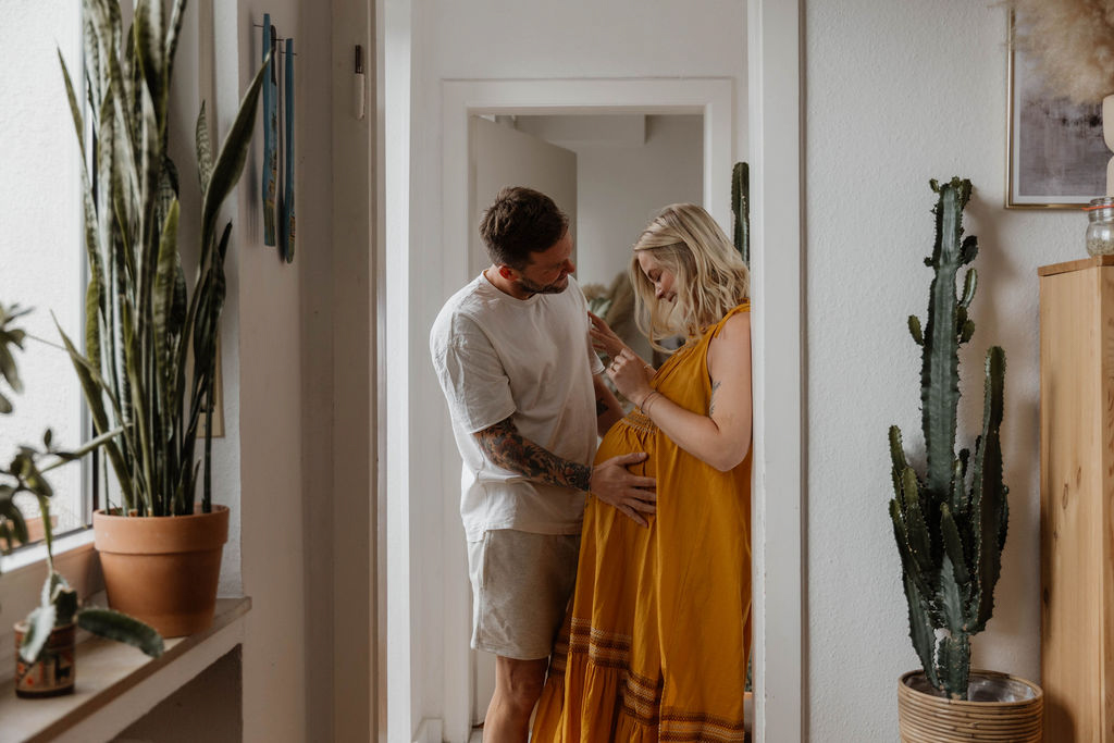 woman leaning against a door frame happily looking down at her pregnant belly her partner standing in front of her hands on her belly