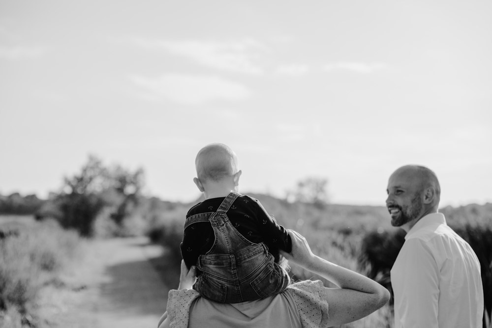 black and white photo family of three photographed from behind child sitting on his mother's shoulders father smiling at them