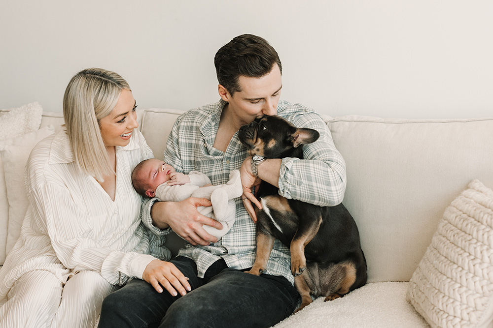 parents sitting on a couch father is holding their newborn while giving their dog a forehead kiss