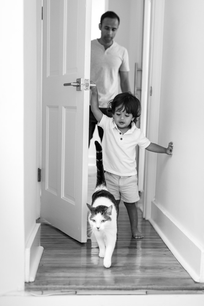 black an white photo of a toddler coming through a door following his white and grey cat both are followed by the father
