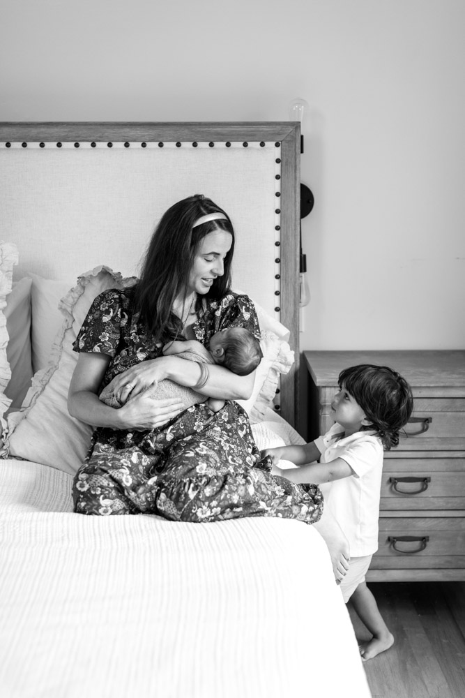black and white photo of mother sitting on the bed holding her newborn laughing at her toddler who is standing next to the bed looking at them