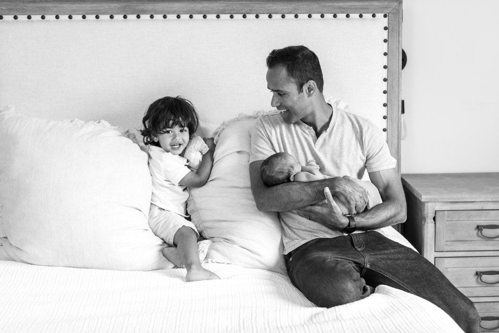 black and white photo of father sitting on the bed holding the newborn in is arms laughing at his toddler who is sitting between pillows smiling at the camera