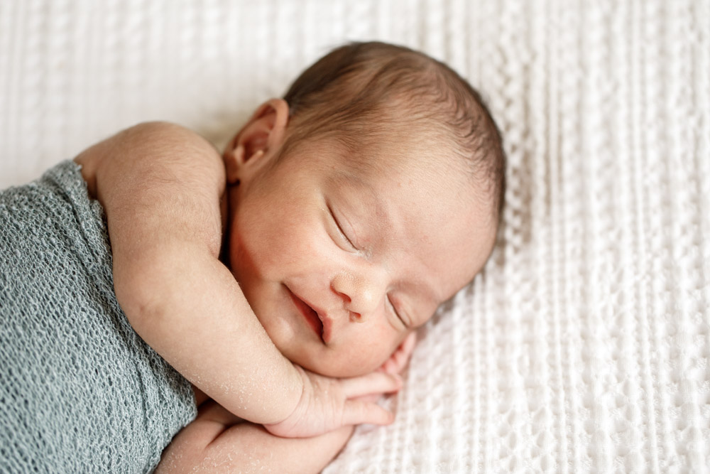 close up of newborn wrapped up in a blanket sleeping on its side with hand tucked under the chin lying on a white blanket smiling