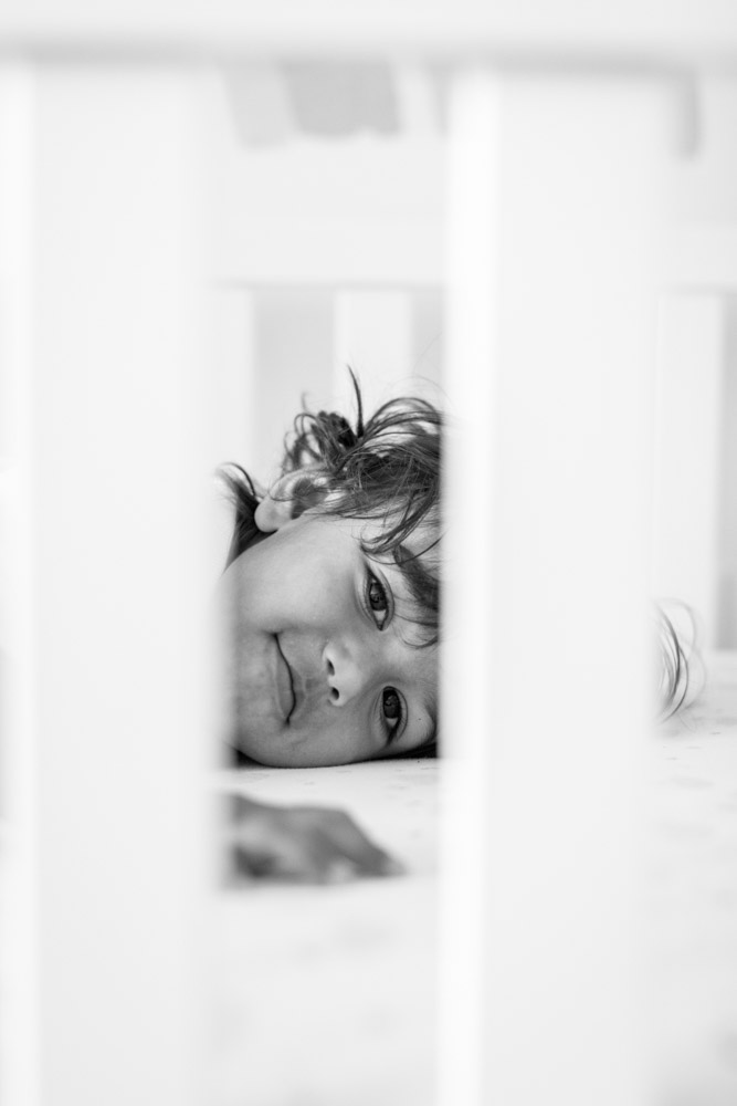 black and white close up of toddler lying in the crib looking through the bed posts smiling at the camera