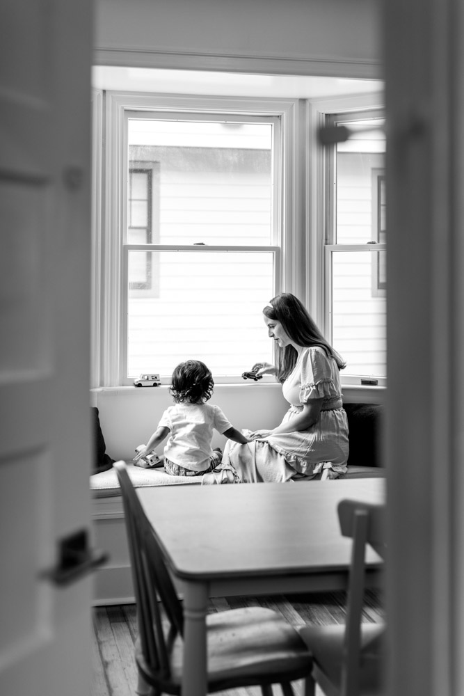 black and white photo of mother and son from behind sitting on a bench in front of the window playing with toy cars while little boy is touching his moms knee