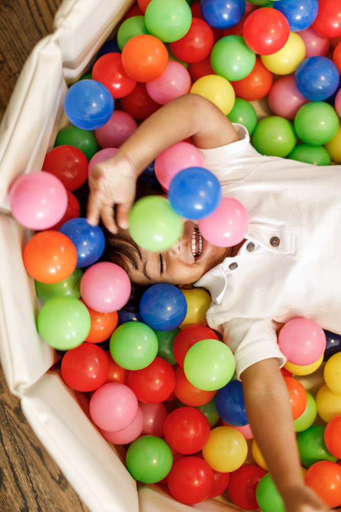 little boy lying in a ball pit full of colorful balls smiling with his left arm up eyes closed