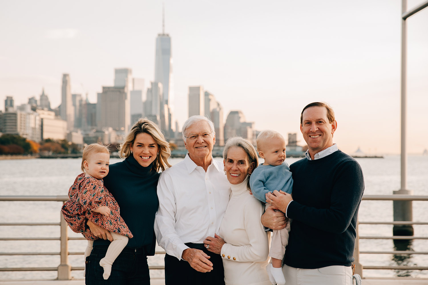 family of six with Manhattan skyline in the background mother holding her little daughter on the left father holding his baby boy on the right and grandparents standing in the middle all smiling at the camera