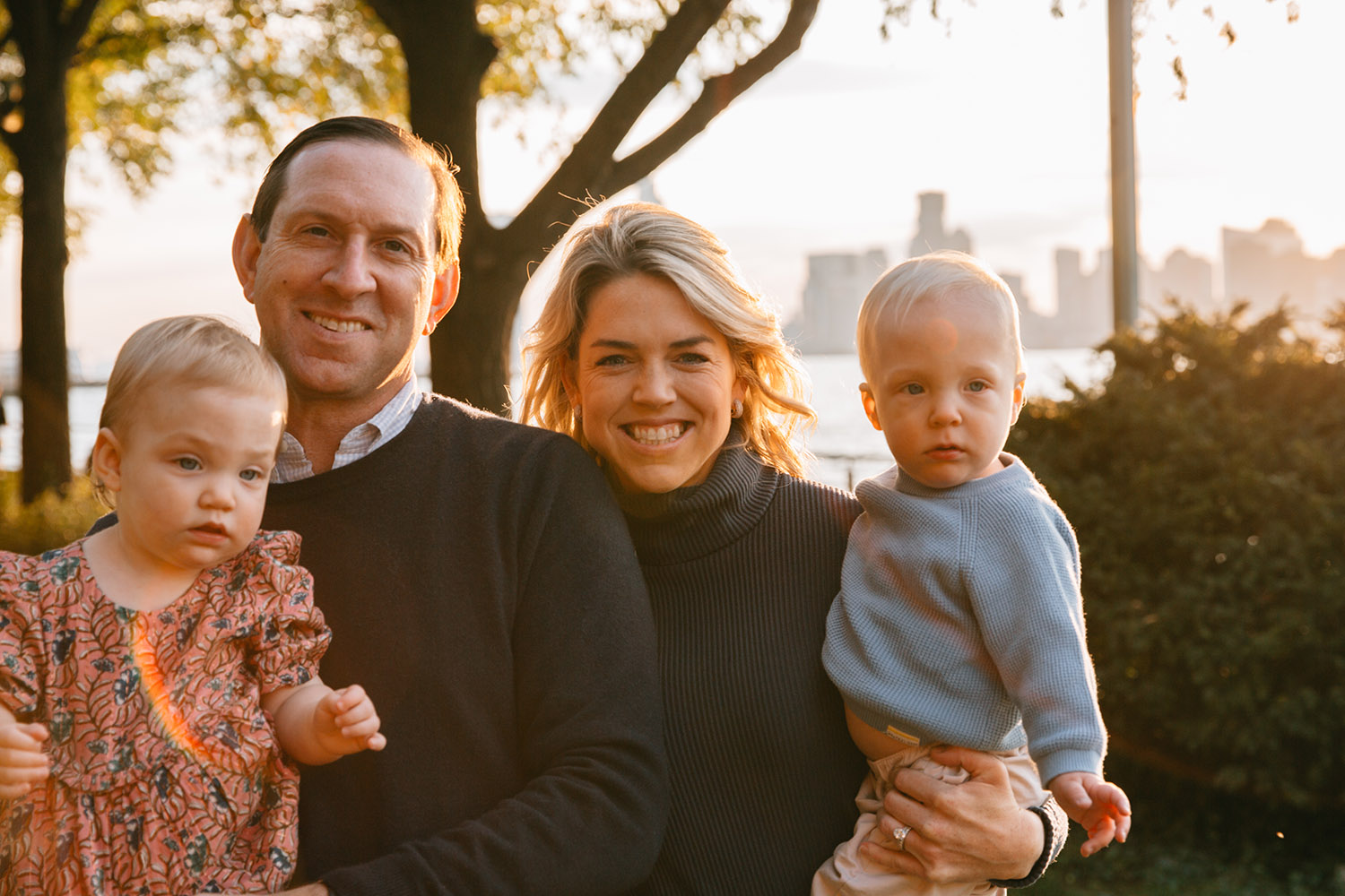 family portrait of four mother and father are holding their two toddlers and smiling at the camera in the sunset with Manhattan backdrop