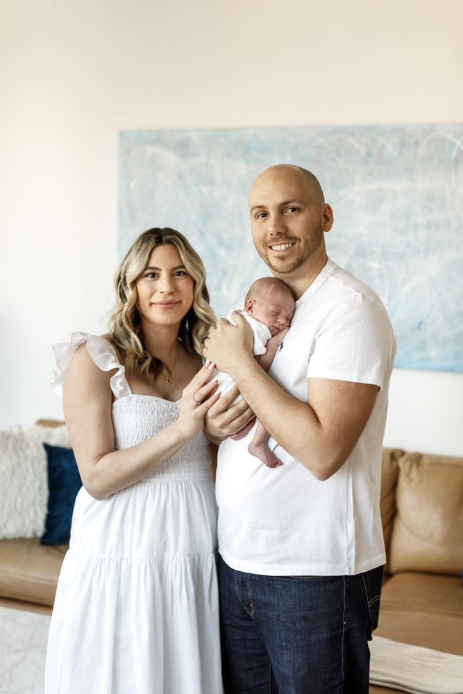 family of three standing in their living room smiling at the camera father is holding the sleeping newborn baby