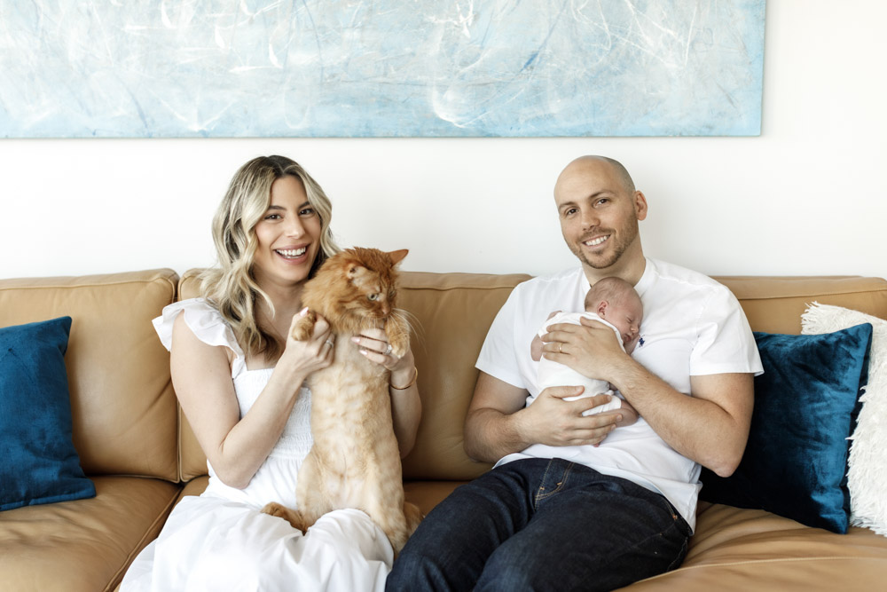 couple sitting in the couch father is holding the sleeping newborn and mother is holding their beautiful brown cat while smiling at the camera