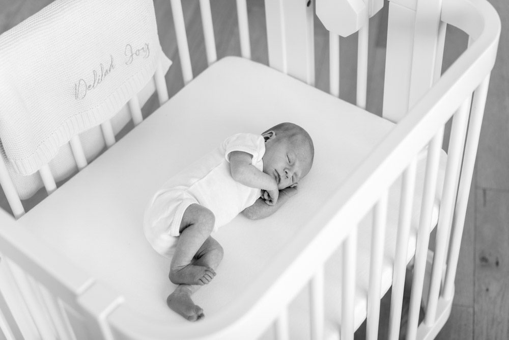 black and white photo of a sleeping newborn lying on its side in the crib with hand tucked under the chin