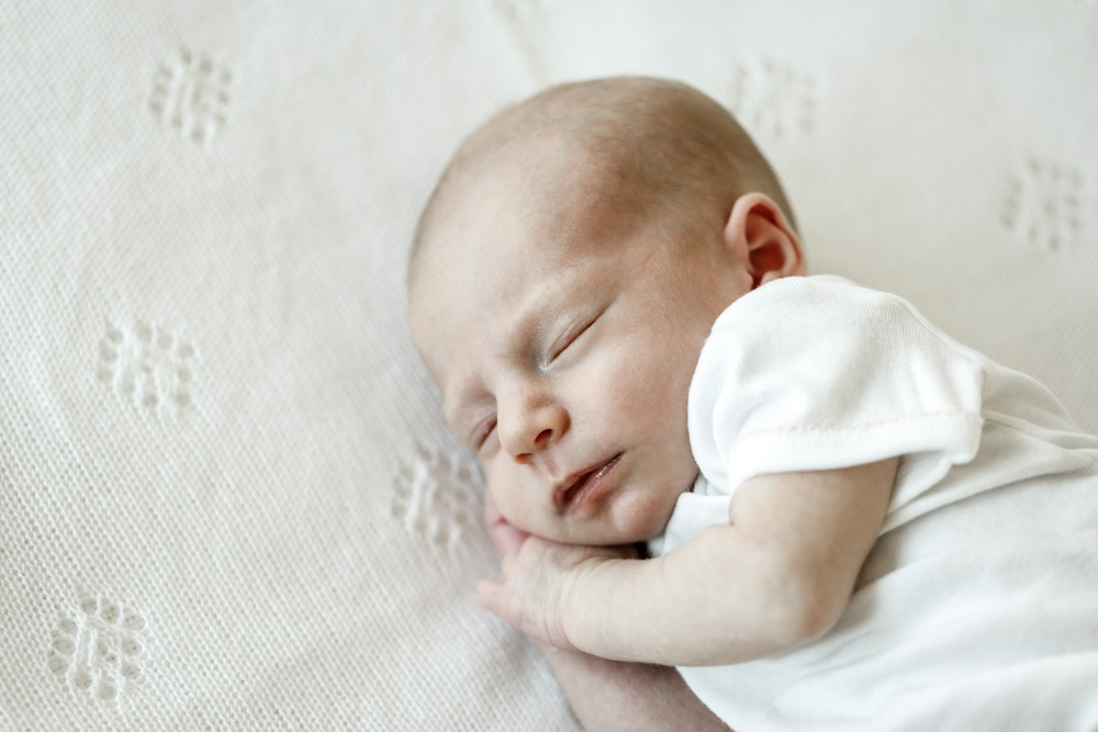 close up of newborn wearing a white onesie sleeping on its side with hand tucked under the chin lying on a beige blanket