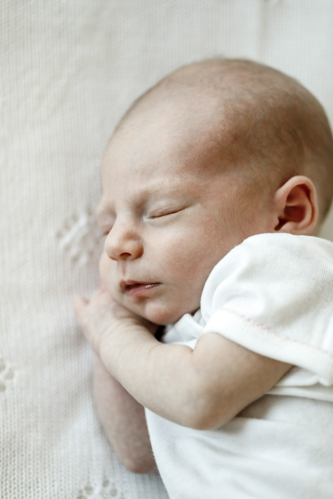close up of newborn wearing a white onesie sleeping on its side with hand tucked under the chin lying on a beige blanket
