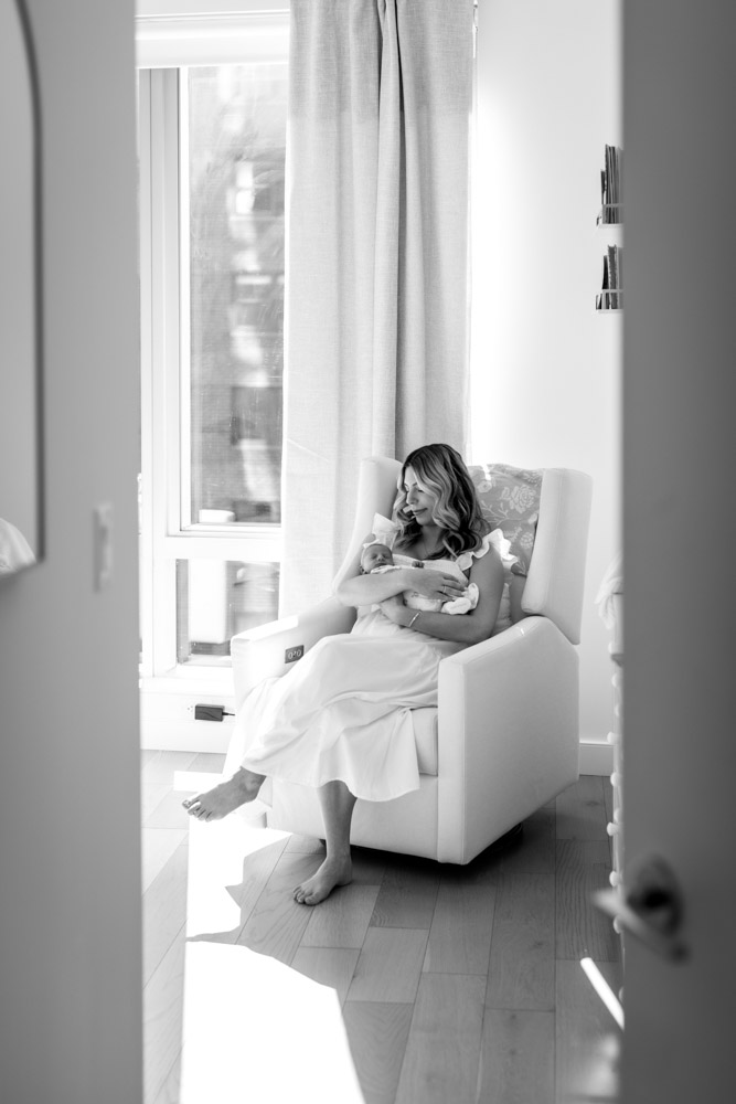 black and white photo of mother in a white dress sitting in a comfortable chair holding her newborn in her arms