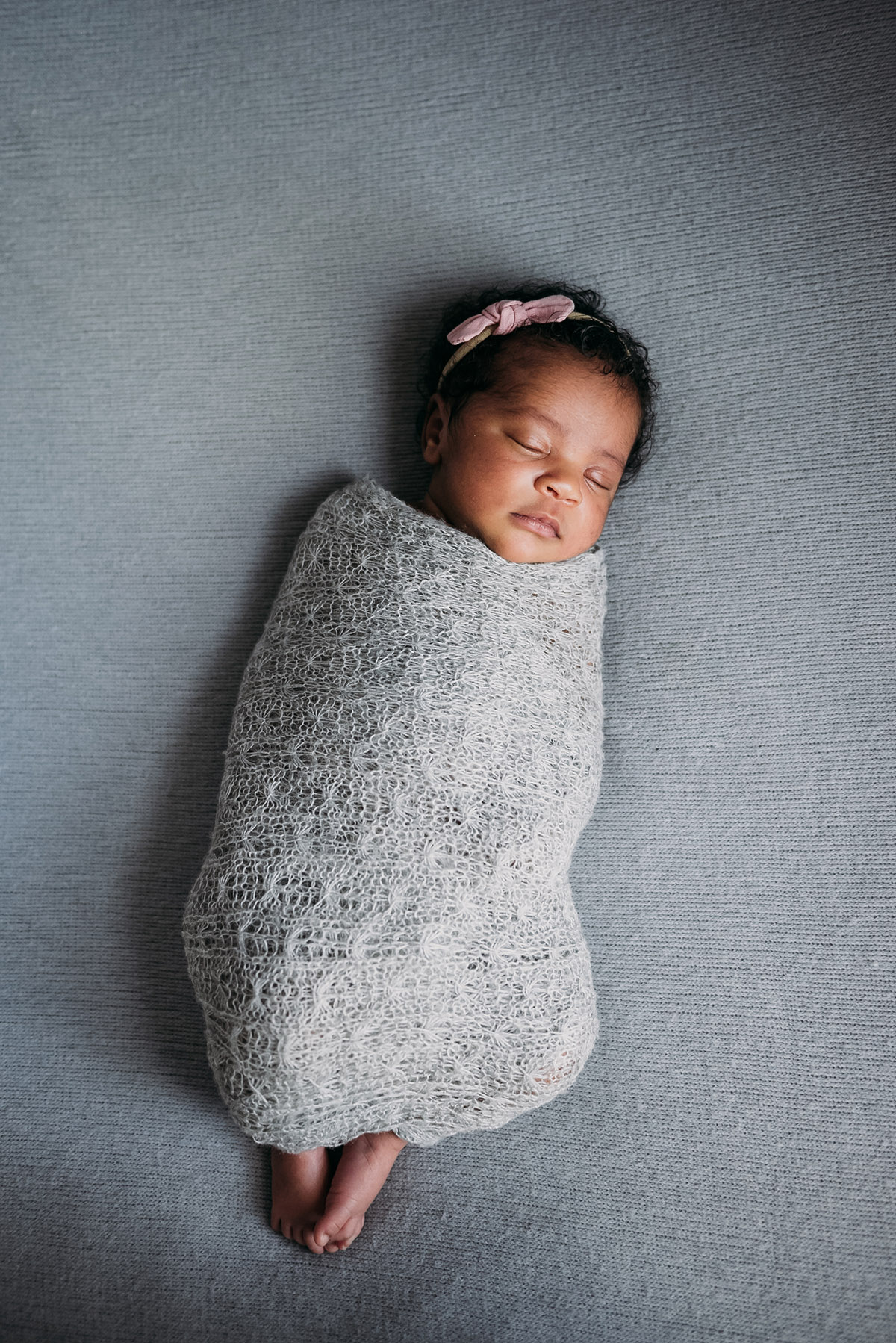 newborn swaddled in a grey blanket with its feet sticking out wearing a headband eyes closed