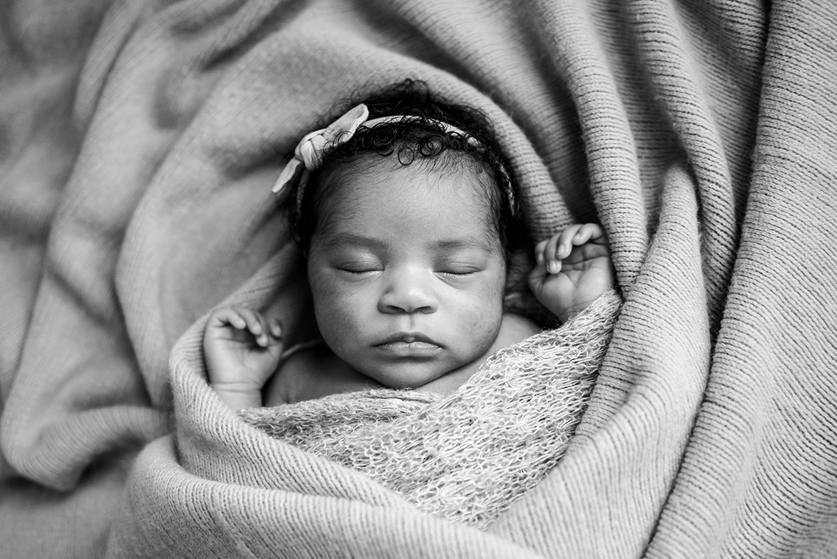black and white photo of a newborn wearing a headband wrapped up in a blanket sleeping on its back with arms sticking out