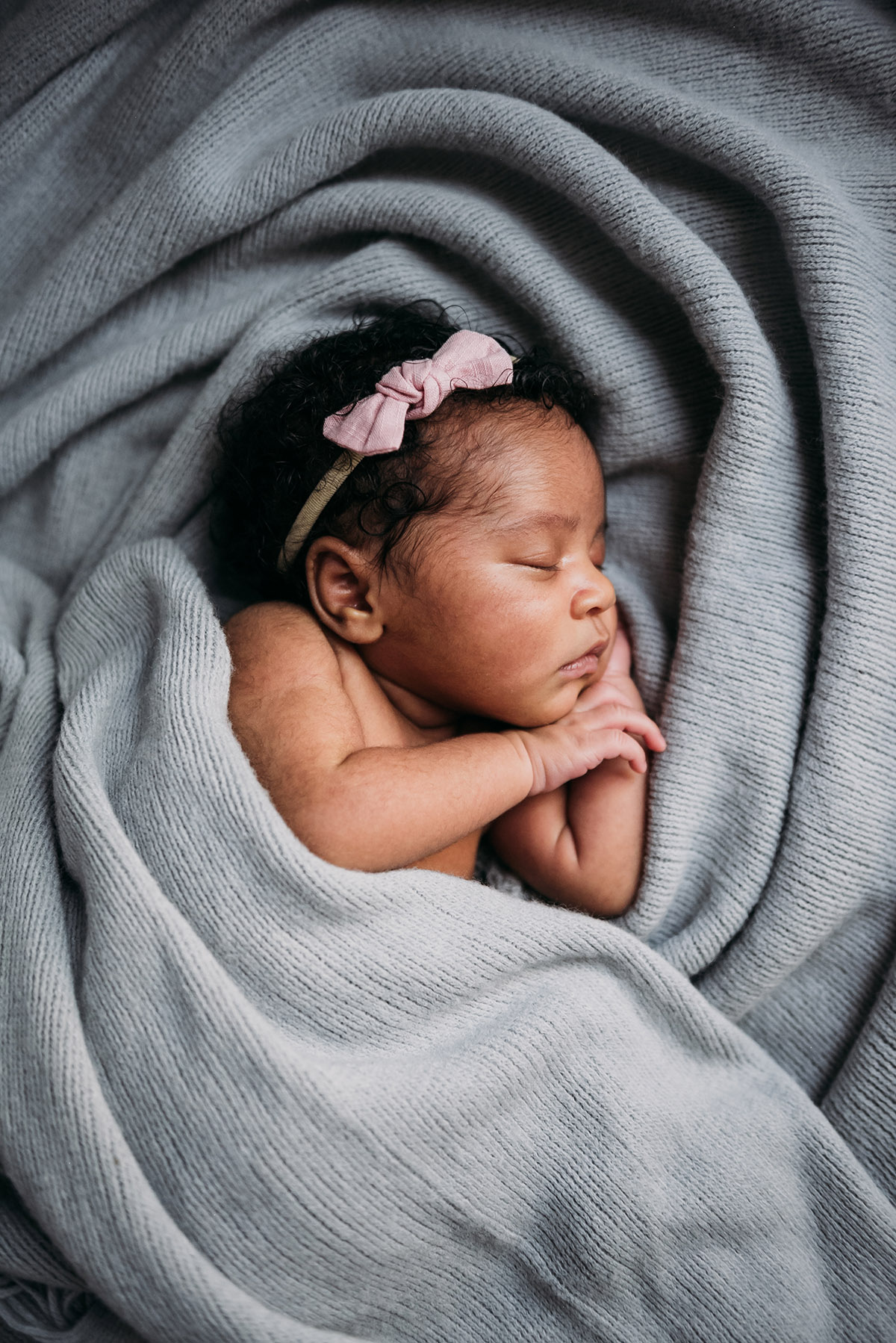 close up of a newborn wearing a headband wrapped up in a grey blanket lying on its side sleeping