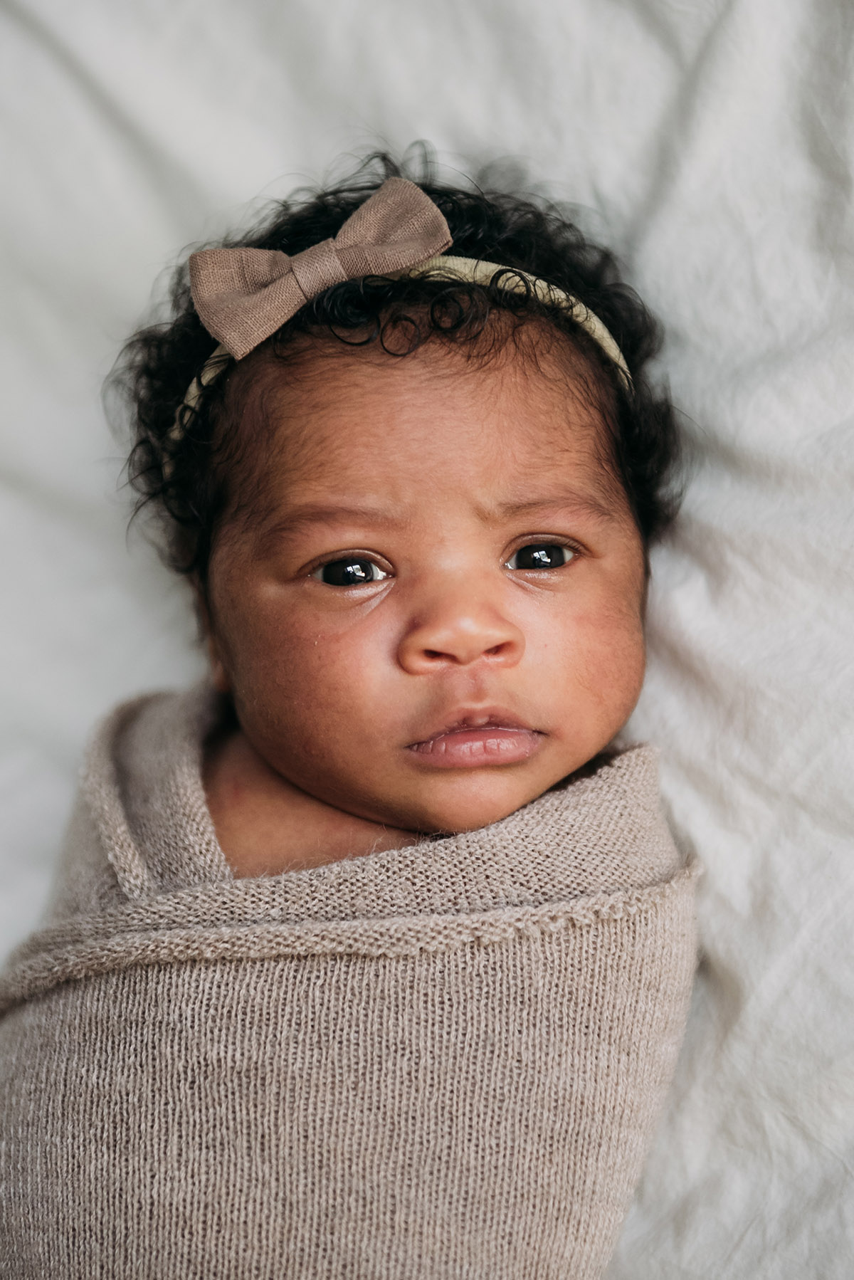 close up of newborn wrapped up in a beige blanket wearing a headband looking at the camera