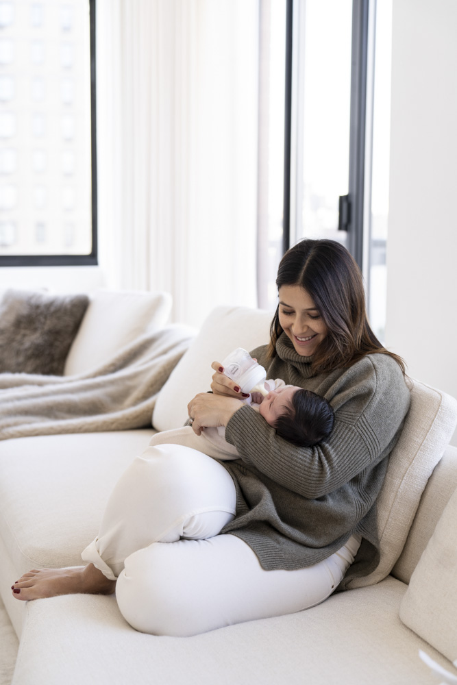 mother sitting on the couch in front of a big window bottle feeding her newborn smiling at it