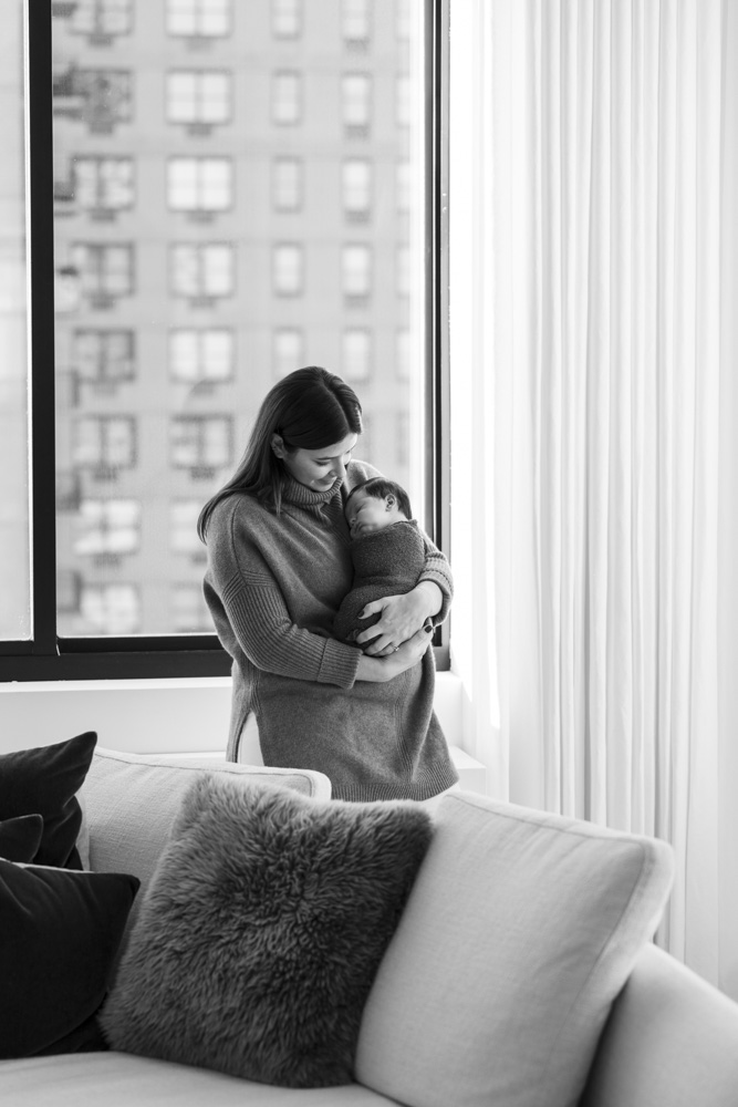 black and white photo of mother holding her newborn standing in front of a big window looking at her baby